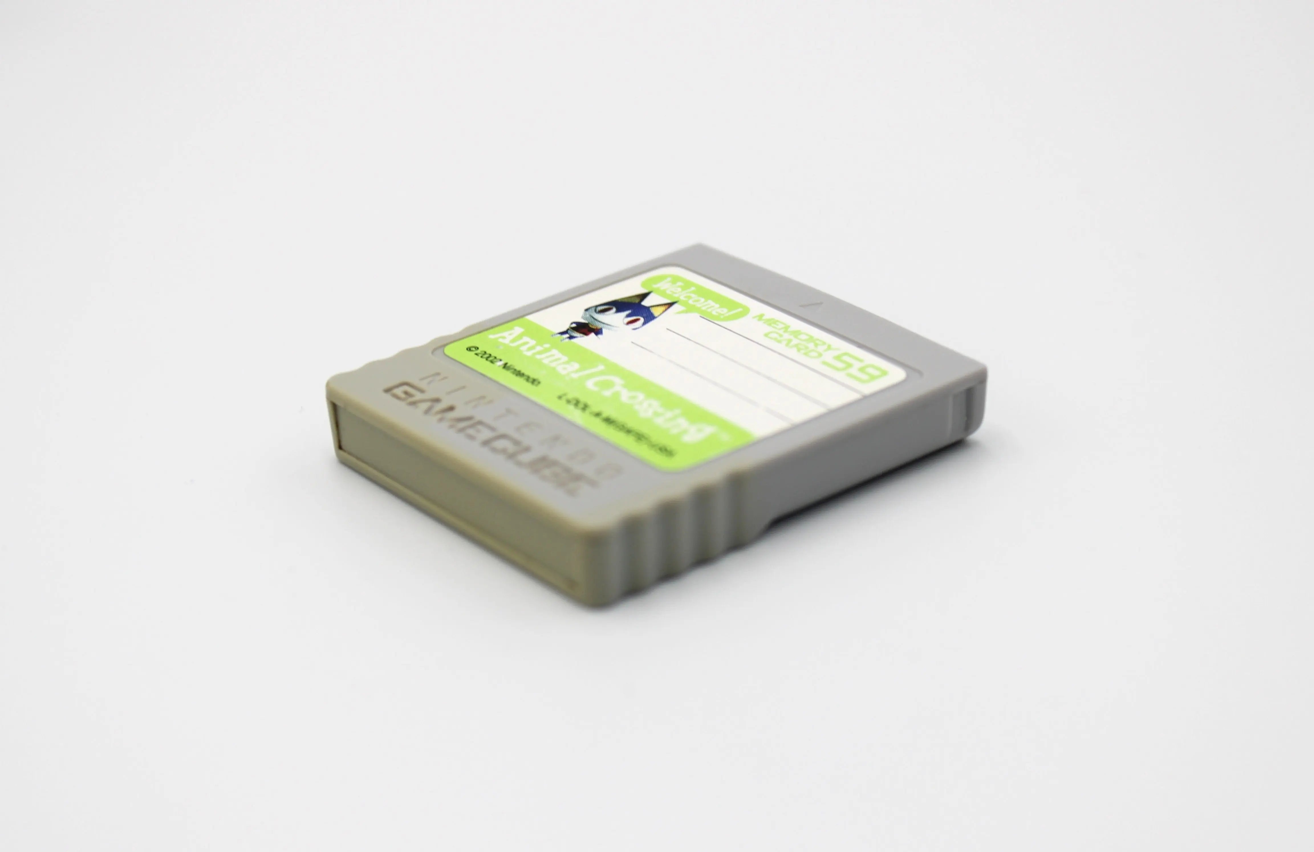 Side angle of front left of cartridge image of 2002 released NTSC-U of OEM Animal Crossing Memory Card for Nintendo GameCube – 59 Blocks (DOL-008). Displaying authentic grey Animal Crossing memory card front at a left angle with intact label and minor discoloration to the bottom of the cartridge on a clean white background.
